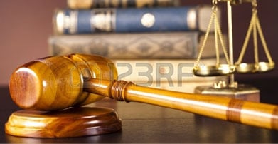 A court gavel on a table, with a scales of justice court scale and law books in the background all suggesting settlement of a case.