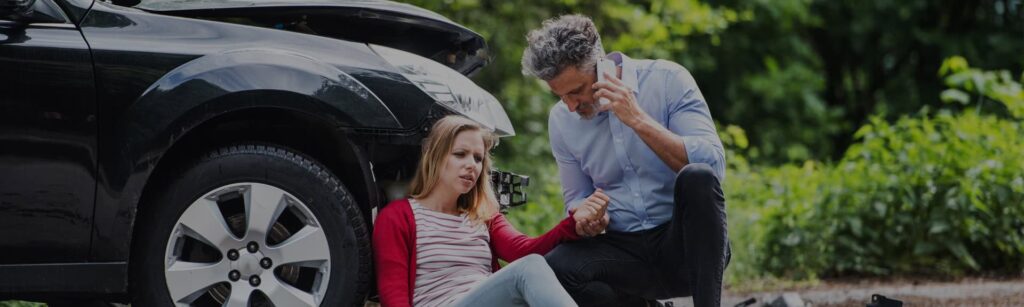 A person is checking on the victim of a car accident right by her car while he calls police.